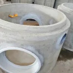 Betonowy krąg studzienny z otworami, widoczny żółty element montażowy, szare tło magazynowe.