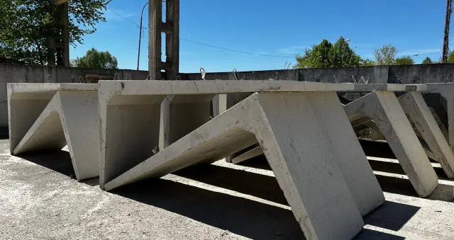 Szereg prefabrykowanych elementów betonowych o specyficznym kształcie litery 'V', prawdopodobnie przeznaczonych do budowy przepustów lub kanalizacji, ułożonych na placu składowym w słoneczny dzień.