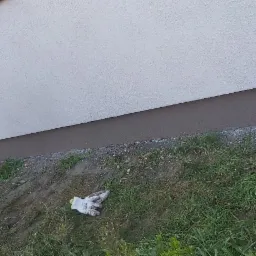 Fragment ocieplonej elewacji budynku z widoczną dolną, pomalowaną na brązowo strefą cokołową, obok leżą zabrudzone rękawice robocze na tle trawy i roślinności.