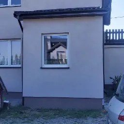 Jasna elewacja domu jednorodzinnego z widoczną przybudówką i oknami, z boku zaparkowany samochód dostawczy, w tle latarnia uliczna.