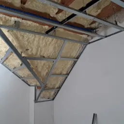 Skośne poddasze w trakcie ocieplania pianą, widoczna metalowa konstrukcja sufitu podwieszanego i niebieskie rurki instalacji elektrycznej.