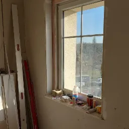 Wnętrze w trakcie montażu płyt kartonowo-gipsowych wokół okna, widoczne profile sufitowe, poziomice opierające się o ścianę i akcesoria na parapecie okiennym.