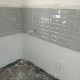 Świeżo ułożone, błyszczące, szare płytki typu metro na ścianie w trakcie remontu łazienki, z widocznymi przewodami elektrycznymi i zabezpieczoną folią podłogą.