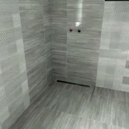 Wykończona kabina prysznicowa z szarymi płytkami o zróżnicowanych wzorach i teksturach, widoczne dwa zawory i odpływ liniowy w podłodze, fragmenty ściany z niedokończonym wykończeniem w górnym rogu.