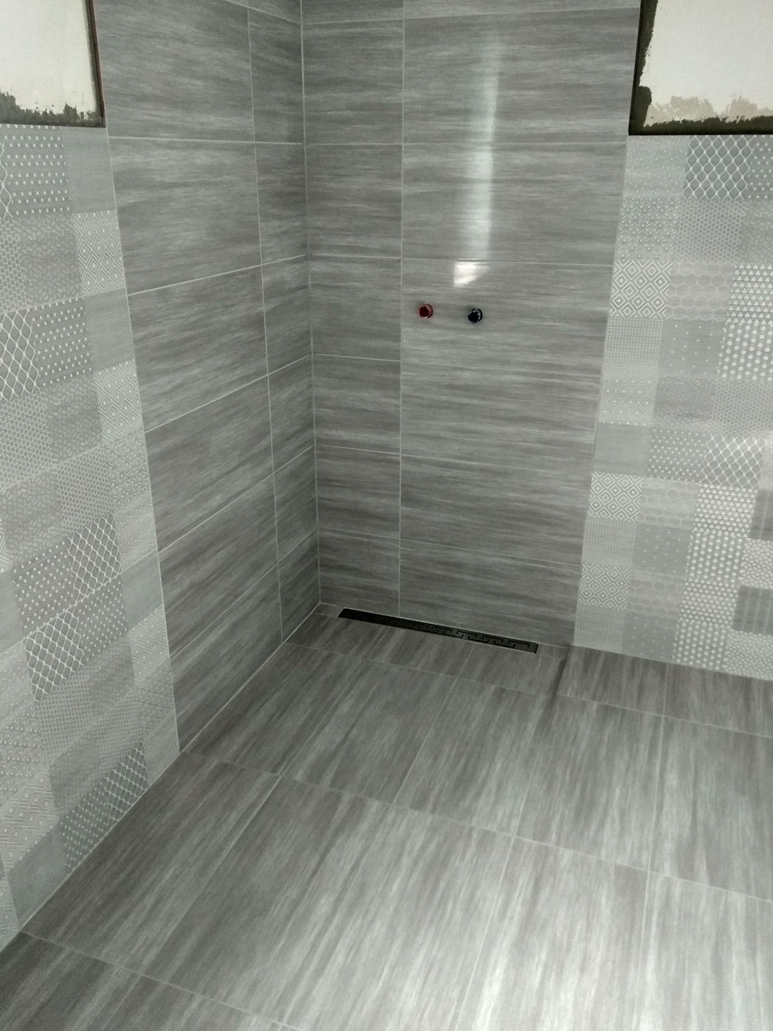 Wykończona kabina prysznicowa z szarymi płytkami o zróżnicowanych wzorach i teksturach, widoczne dwa zawory i odpływ liniowy w podłodze, fragmenty ściany z niedokończonym wykończeniem w górnym rogu.