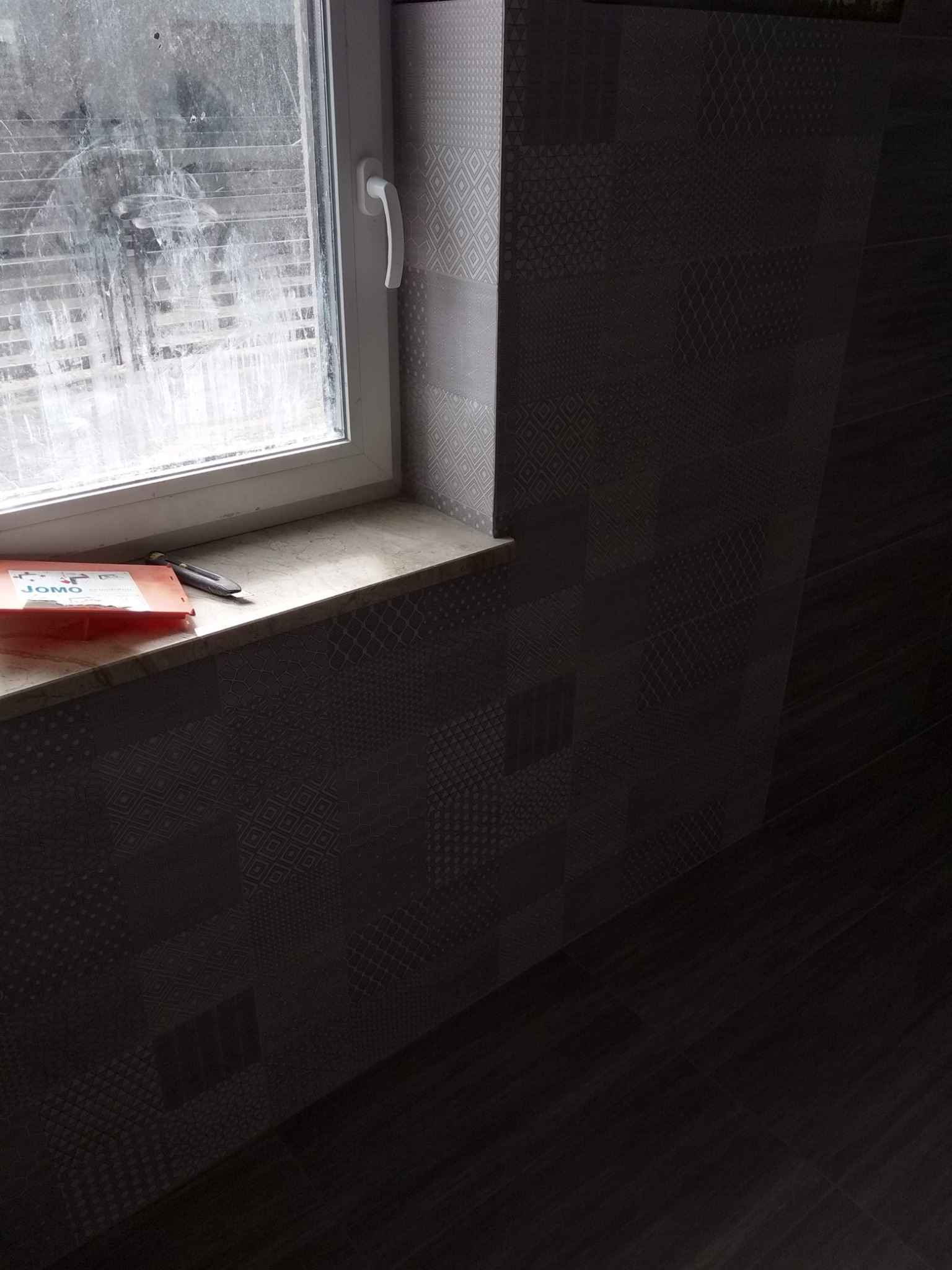 Ułożone ciemne płytki ceramiczne z wzorem na ścianie i podłodze, widoczny parapet z narzędziami, okno brudne.