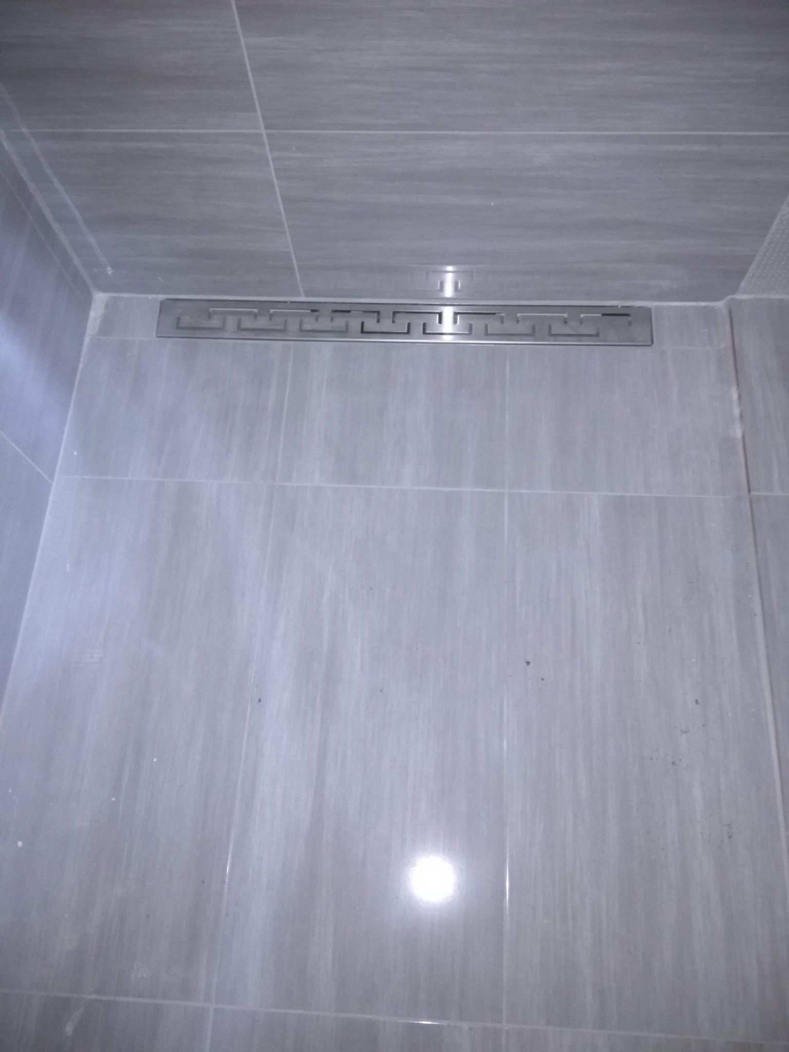 Szare płytki w kabinie prysznicowej z odpływem liniowym ozdobionym wzorem greckim.