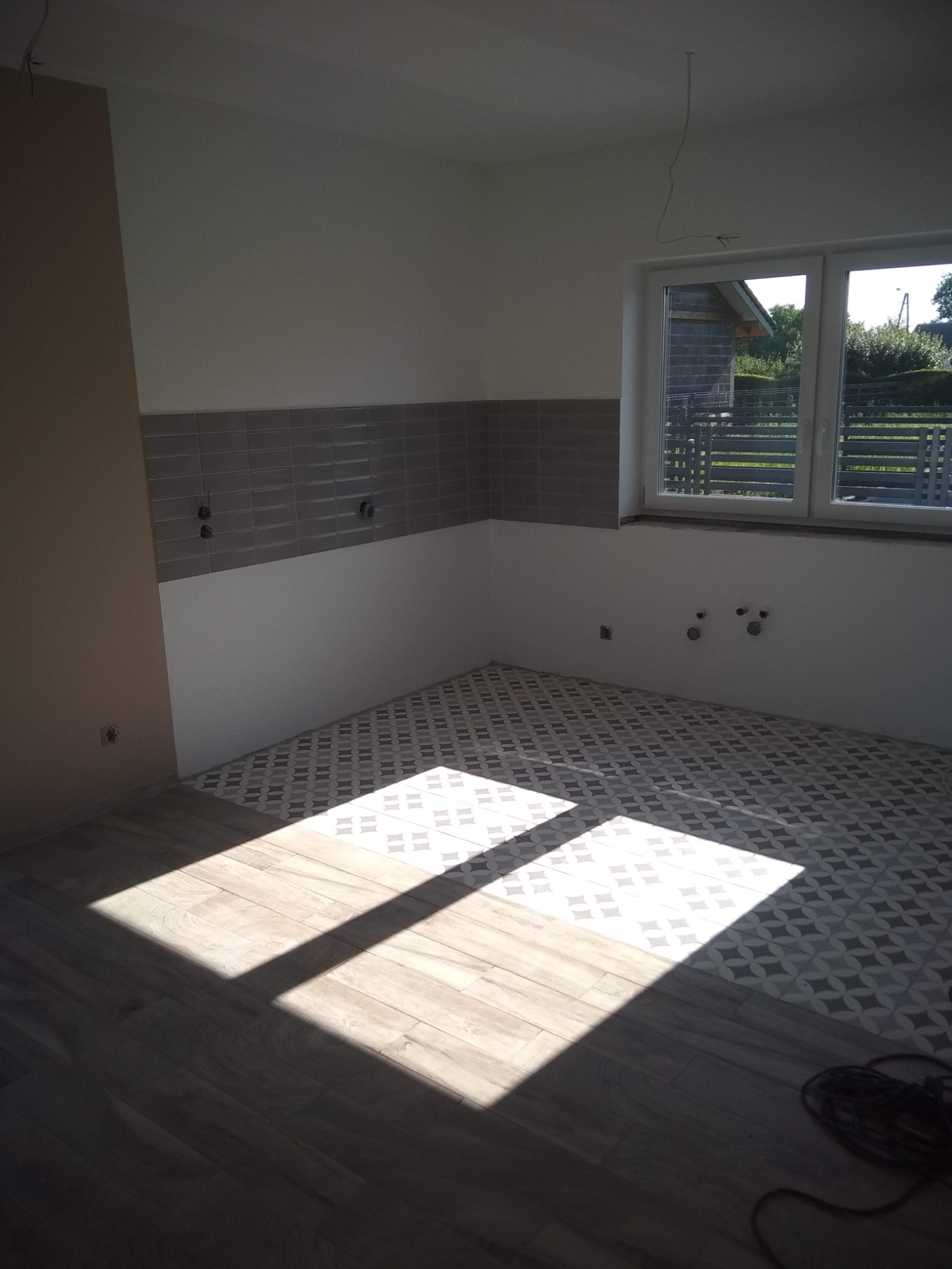 Wnętrze kuchni w trakcie wykańczania: ułożona podłoga z płytek w geometryczny wzór oraz imitująca drewno, szare płytki na ścianie, widoczne wyprowadzenia instalacji hydraulicznej i elektrycznej...