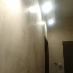 Ściana w korytarzu pokryta dekoracyjną, beżową farbą z efektem przetarć, widoczne oświetlenie sufitowe i fragment ciemnych drzwi.