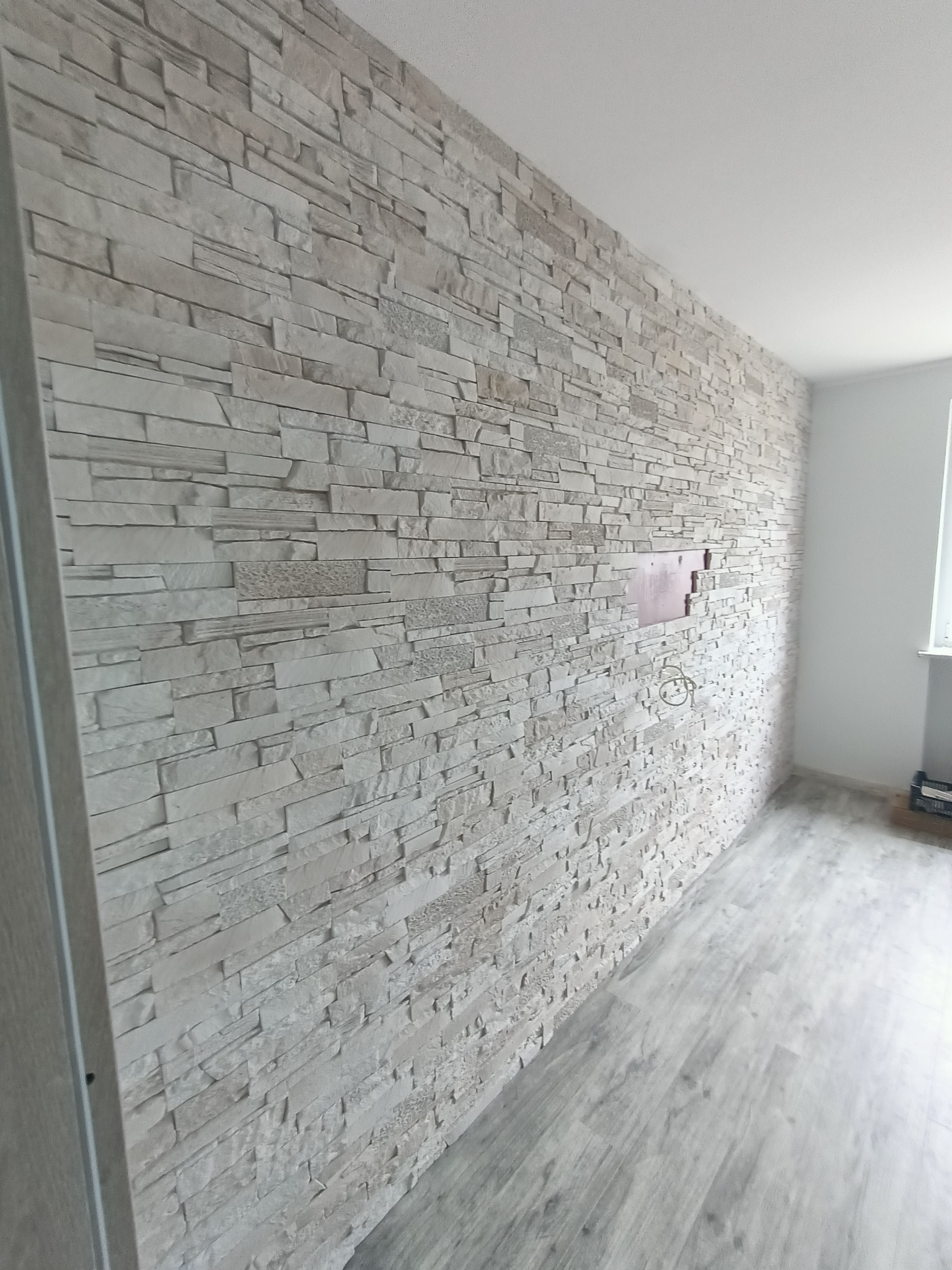 Ściana wykończona dekoracyjną okładziną imitującą kamień, z widocznym otworem na instalację elektryczną i częściowo odsłoniętą ścianą pod okładziną, oraz panelami podłogowymi w odcieniach szarości.