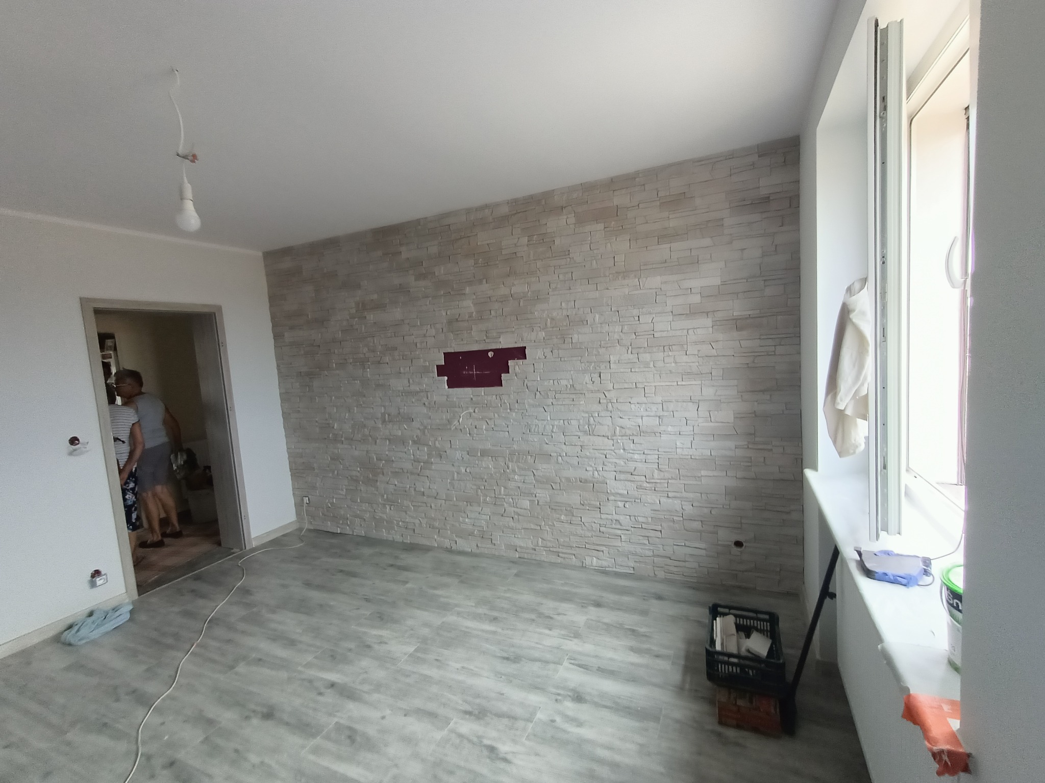 Pokój po remoncie z wykończoną ścianą z dekoracyjnej cegły, widoczne ślady po instalacji, podłoga wyłożona panelami imitującymi drewno, niedokończone oświetlenie sufitowe, okno z resztkami farby...