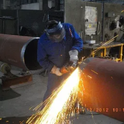 Pracownik w niebieskim kombinezonie i ochronnej masce szlifuje spaw na dużej, pomalowanej rurze za pomocą szlifierki kątowej, iskry rozpraszają się wokół