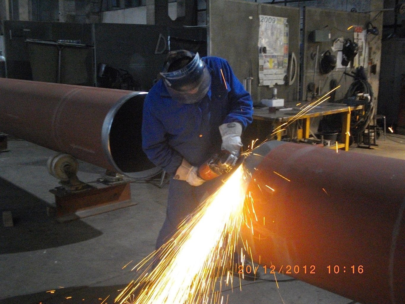 Pracownik w niebieskim kombinezonie i ochronnej masce szlifuje spaw na dużej, pomalowanej rurze za pomocą szlifierki kątowej, iskry rozpraszają się wokół