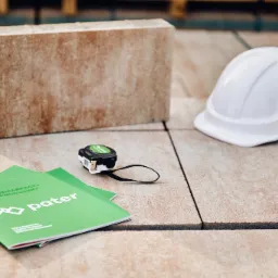 Prezentacja materiałów budowlanych: próbka produktu o porowatej fakturze, katalog firmy 'Pater' w odcieniach zieleni, miara zwijana i biały kask ochronny na ułożonej nawierzchni.