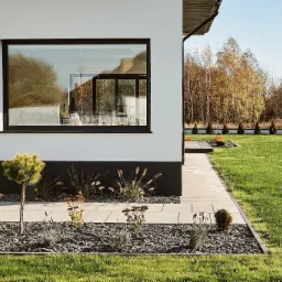 Elegancki chodnik z jasnych płyt prowadzący wzdłuż nowoczesnego domu z białymi ścianami i czarnym cokołem, rabata wysypana korą z ozdobnymi roślinami