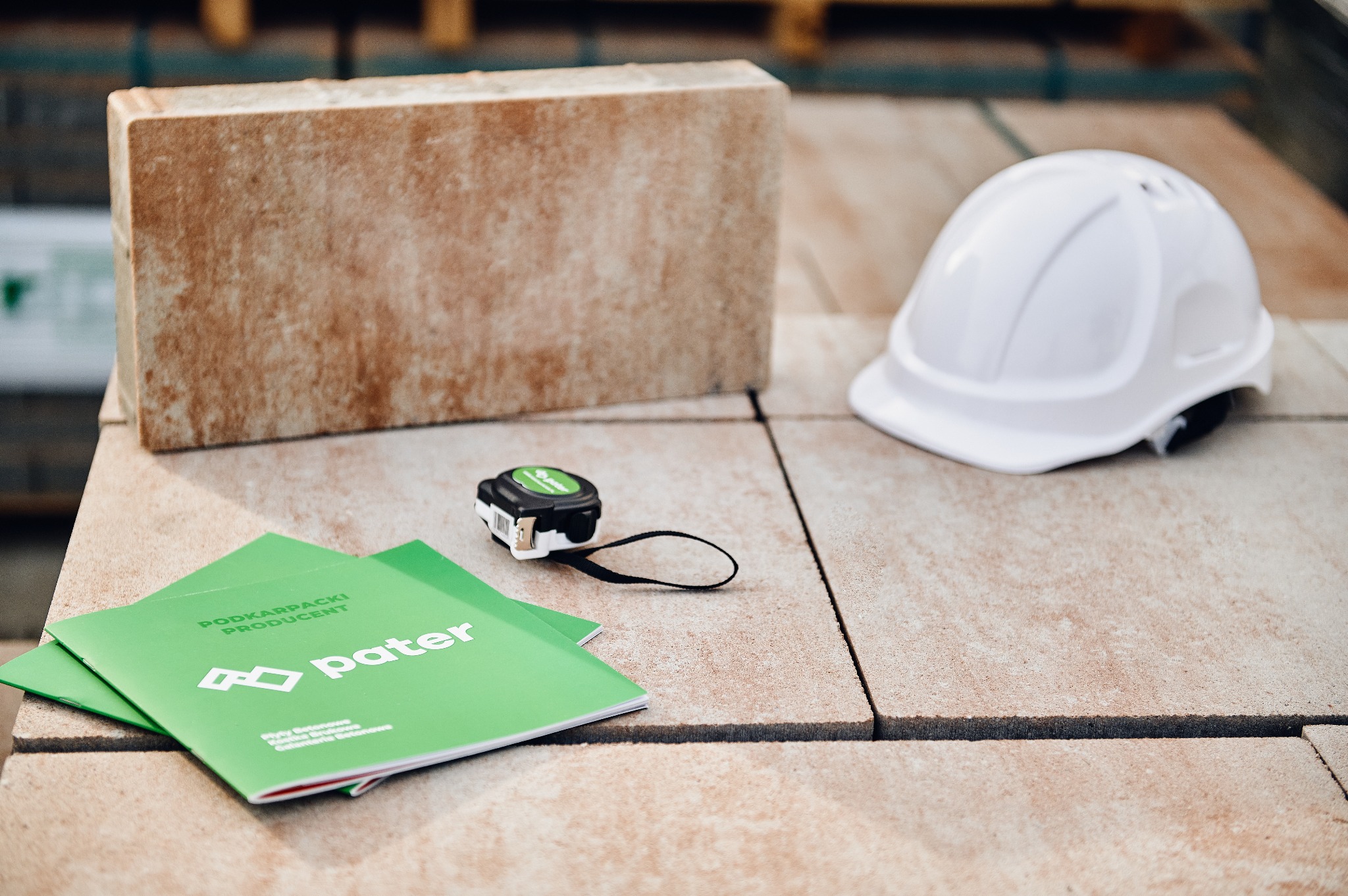 Prezentacja materiałów budowlanych: próbka produktu o porowatej fakturze, katalog firmy 'Pater' w odcieniach zieleni, miara zwijana i biały kask ochronny na ułożonej nawierzchni.