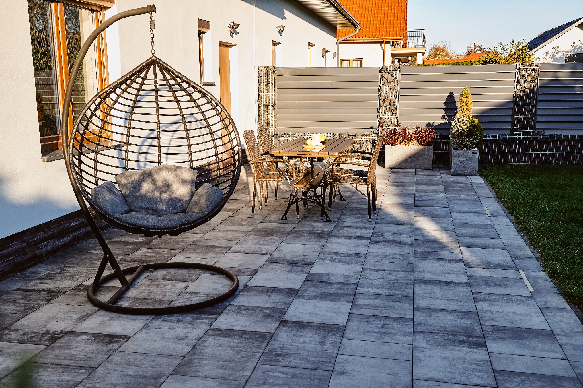 Zewnętrzna przestrzeń z szarymi płytami nawierzchniowymi, wiszącym fotelem z poduszkami, stołem z krzesłami i ogrodzeniem z gabionów oraz roślinami w donicach.