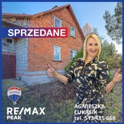 Agnieszka Łukasik Nieruchomości - Agentka nieruchomości RE/MAX Peak na tle sprzedanego, ceglanego domu z napisem 'SPRZEDANE', uśmiechnięta, z uniesionymi kciukami.