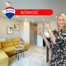 Jasna kawalerka z żółtą sofą i marmurową podłogą prezentowana przez agentkę nieruchomości, z balonem logo RE/MAX i napisem 'NOWOŚĆ'.
