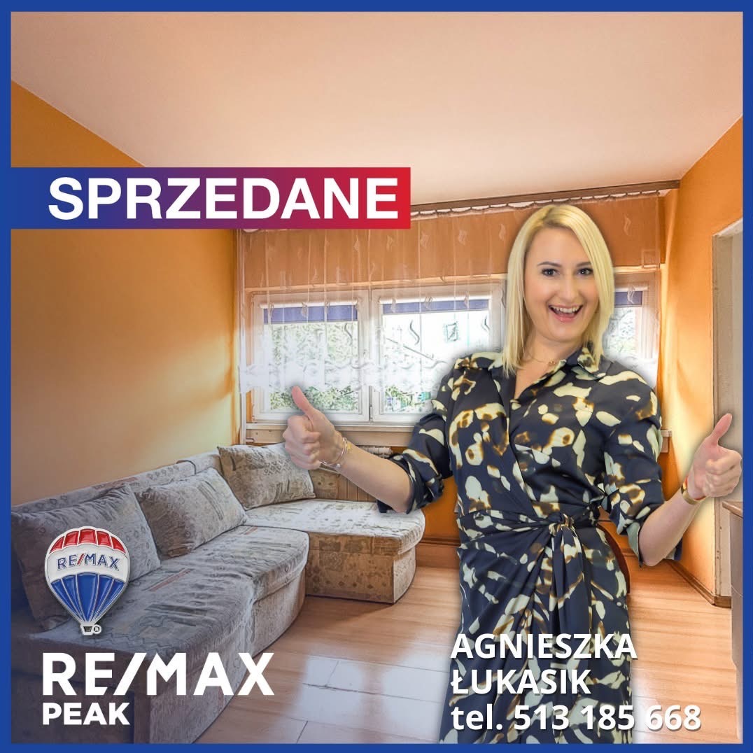 Wnętrze salonu z agentką RE/MAX, uśmiechniętą i pokazującą kciuki w górę. Na górze napis SPRZEDANE. W tle okno z firankami i kanapa.