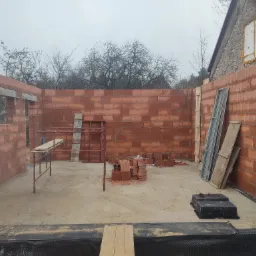 Wnętrze w trakcie budowy domu z czerwonej cegły, widoczne rusztowanie, ułożone cegły, czarna folia izolacyjna na podłożu i fragment ściany sąsiedniego budynku z oknem zabezpieczonym styropianem.