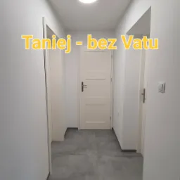 Korytarz po remoncie z szarymi płytkami podłogowymi, białymi ścianami i drzwiami. Na ścianie widoczny włącznik światła. Na zdjęciu widoczny napis 'Taniej - bez Vatu'.