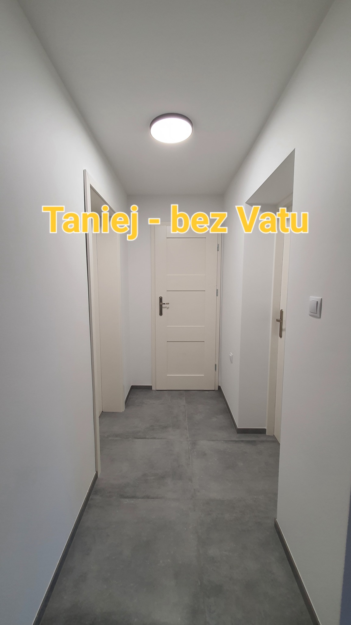 Korytarz po remoncie z szarymi płytkami podłogowymi, białymi ścianami i drzwiami. Na ścianie widoczny włącznik światła. Na zdjęciu widoczny napis 'Taniej - bez Vatu'.
