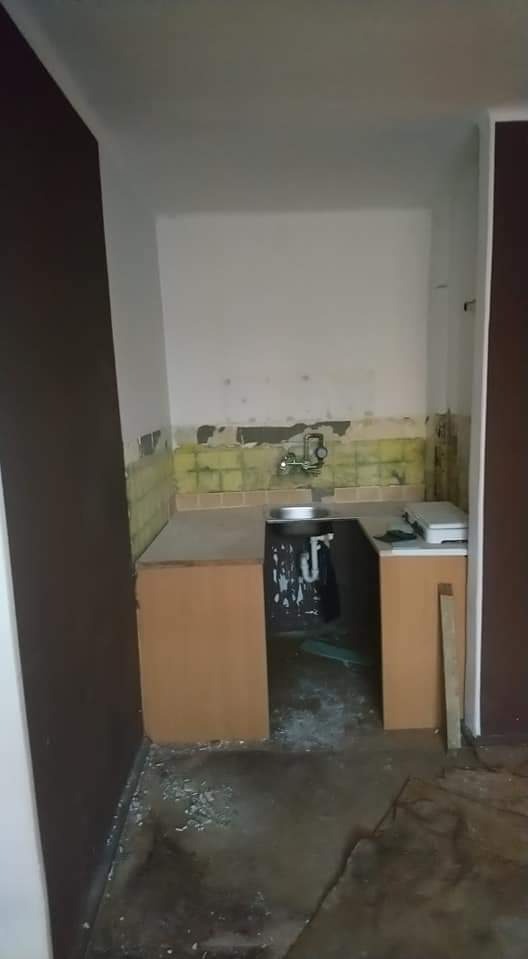 Kuchenny zlew i fragment blatu z szafkami przed remontem: widoczne resztki płytek na ścianie, odpadający tynk, brudna podłoga i elementy wyposażenia przygotowane do wymiany.