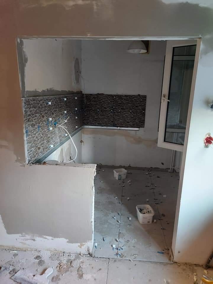 Fragment kuchni w trakcie prac wykończeniowych: częściowo położone ciemne płytki na ścianie, plastikowe krzyżyki dystansowe na podłodze, surowy tynk i niedokończone ściany.