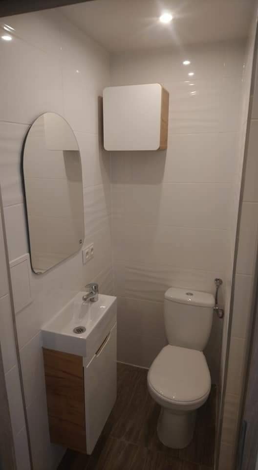 Nowoczesna, mała łazienka z białą armaturą, podwieszaną szafką z umywalką i lustrem w kształcie łuku, wykończona białymi płytkami i brązowymi płytkami podłogowymi imitującymi drewno.