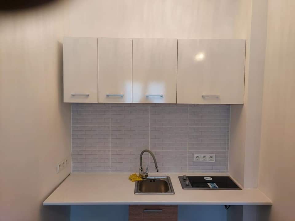 Nowoczesna, jasna kuchnia z białymi szafkami wiszącymi, szarą mozaiką nad blatem, stalowym zlewozmywakiem i płytą indukcyjną, wykończona z dbałością o detale.