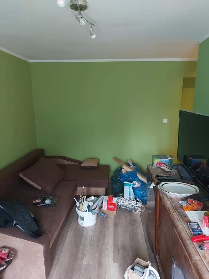 Pokój z zielonymi ścianami w trakcie malowania; widoczny brązowy narożnik, wiadro z pędzlami i wałkami, oraz zabezpieczona folią podłoga z panelami. Część mebli przesunięta, widoczny bałagan.