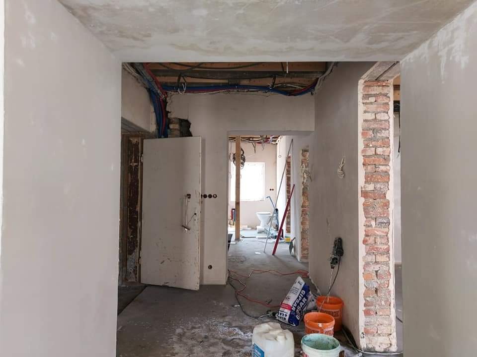 Wnętrze w trakcie prac wykończeniowych: widoczne odkryte instalacje pod sufitem, otynkowane ściany, fragment ściany z cegły, wiaderka z zaprawą i worki z gipsem na podłodze, w tle toaleta.