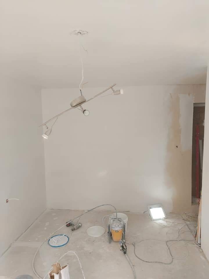 Pokój w trakcie malowania natryskowego na biało, widoczny agregat malarski, lampa oświetleniowa i wisząca lampa sufitowa.