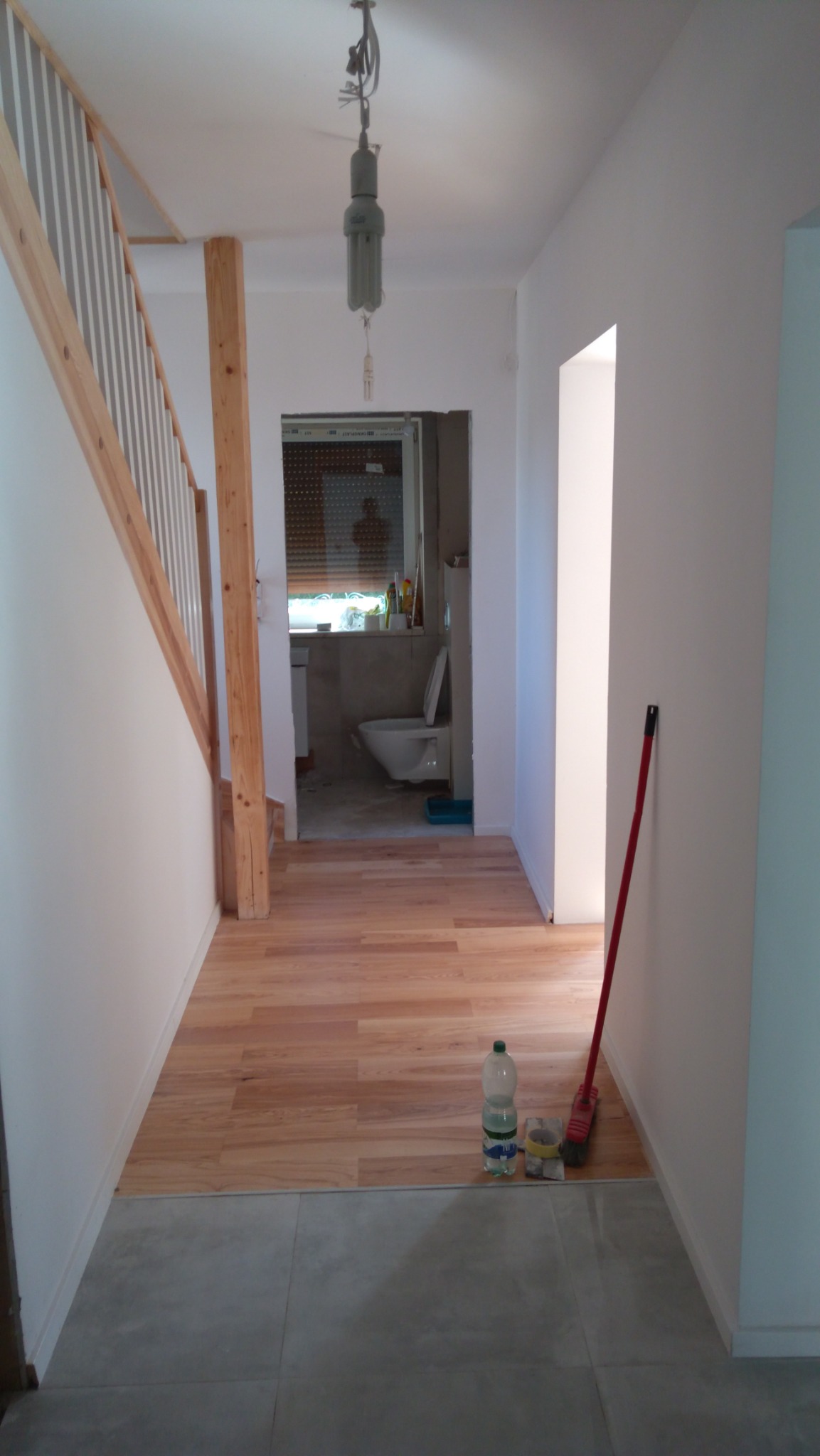 Korytarz w trakcie remontu: ułożone panele podłogowe z widocznym przejściem do łazienki i schodami, butelka wody i narzędzia na podłodze, surowa żarówka na kablu.