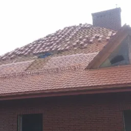 Ceglasty dach karpiówka w trakcie układania, widoczne fragmenty łat i kontrłat, komin z cegły, budynek z czerwonej cegły, częściowo zasłonięty pniem drzewa.