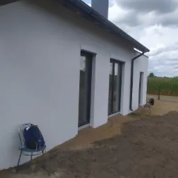 Fragment otynkowanej elewacji domu jednorodzinnego z ciemnymi ramami okiennymi, widoczny plecak na krześle i świeżo wyrównany teren wokół budynku.