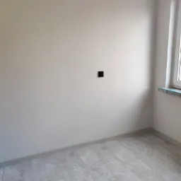 Pomalowane na jasno szaro pomieszczenie z szarymi płytkami podłogowymi, widoczny fragment okna z marmurowym parapetem i czarne gniazdko elektryczne na ścianie.