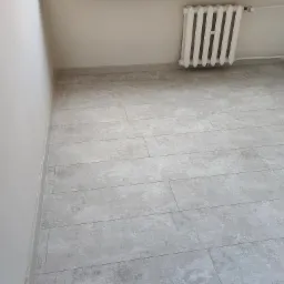 Jasnoszara podłoga z paneli ułożonych wzdłuż ściany, widoczny biały grzejnik żeberkowy i fragment białej ściany z listwą przypodłogową.