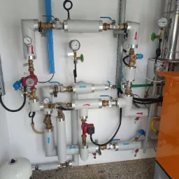 Rozbudowana instalacja grzewcza z piecem gazowym, widoczne rury, pompy obiegowe, manometry i zawory, strzałki wskazują kierunek przepływu, całość zamontowana na białej ścianie.