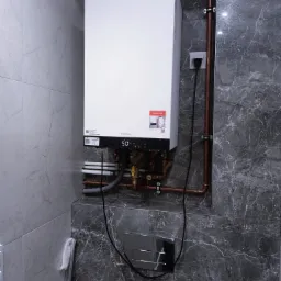 Biały, wiszący piec gazowy Viessmann zamontowany na marmurowej ścianie, z widocznymi rurami miedzianymi i elementami instalacji. Nowoczesne wykończenie łazienki.