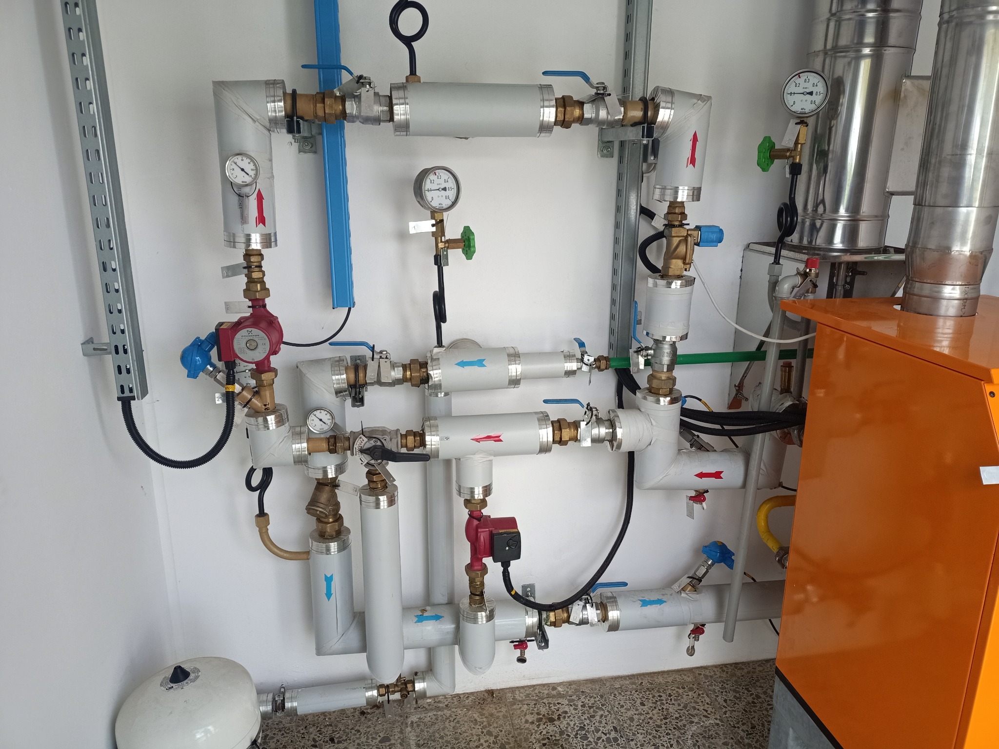 Rozbudowana instalacja grzewcza z piecem gazowym, widoczne rury, pompy obiegowe, manometry i zawory, strzałki wskazują kierunek przepływu, całość zamontowana na białej ścianie.