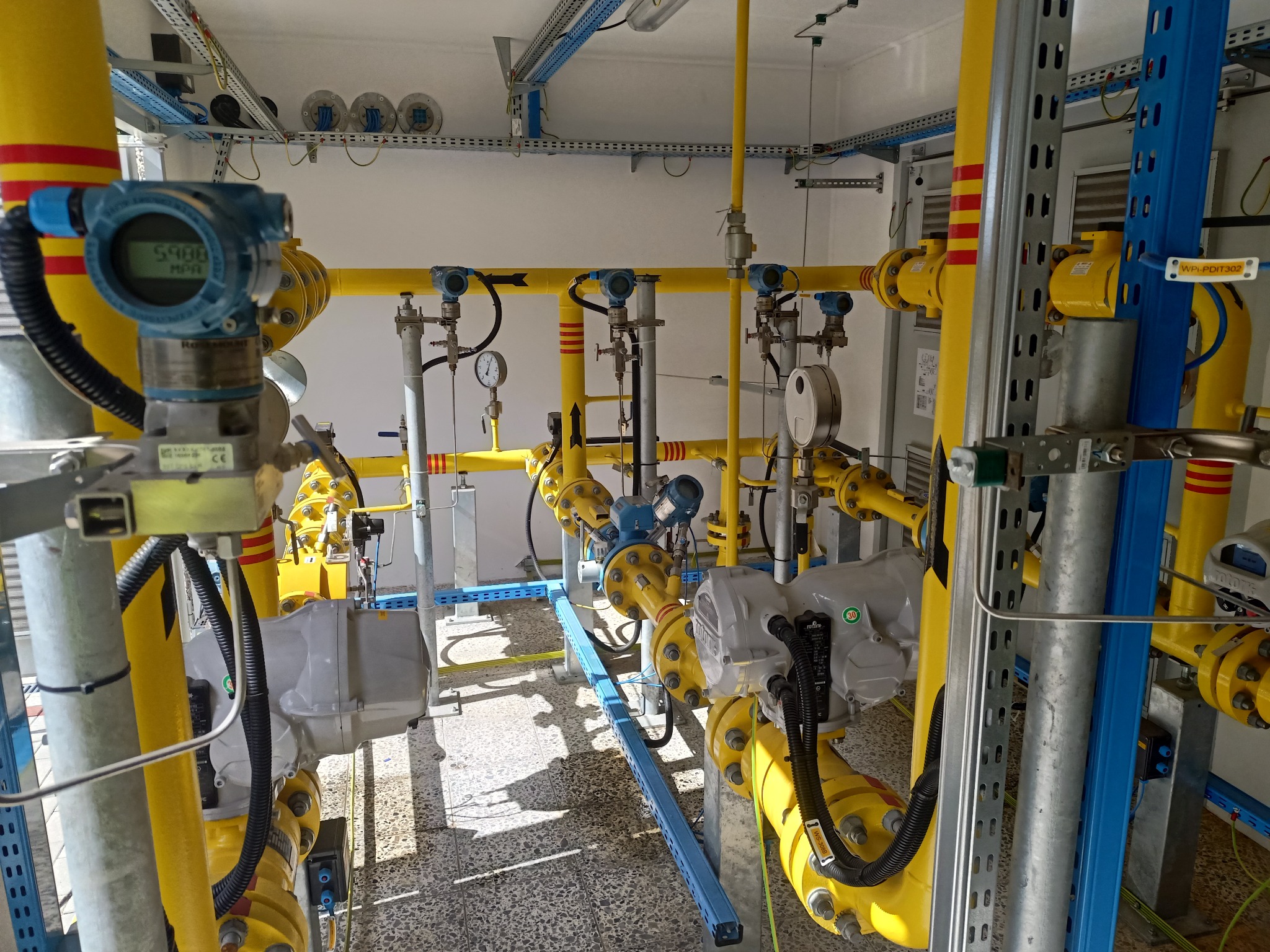 Kompleksowy system rur gazowych w kolorze żółtym z czerwonymi paskami, wyposażony w czujniki ciśnienia i zawory, zamontowany wewnątrz budynku z szarą podłogą i białymi ścianami.