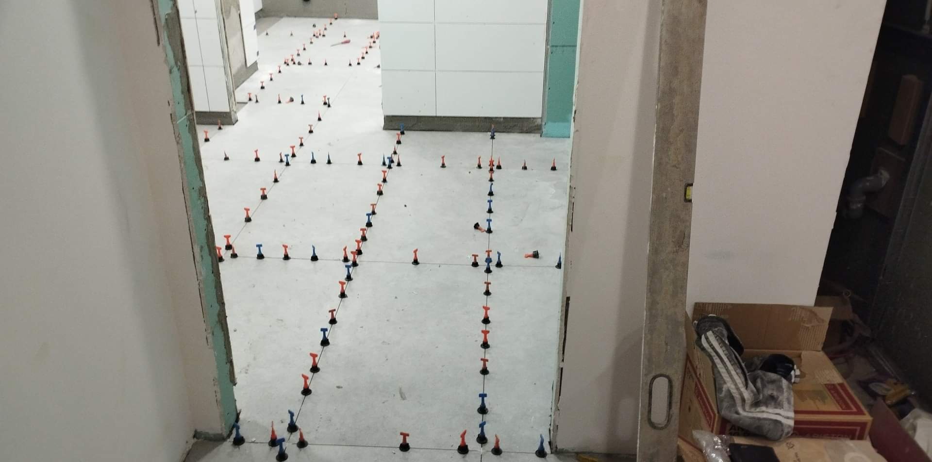 Przygotowanie podłoża pod płytki: widoczne krzyżyki dystansowe w kolorach czerwonym i niebieskim, wyznaczające linie układania w pomieszczeniu z częściowo położonymi płytkami na ścianach.