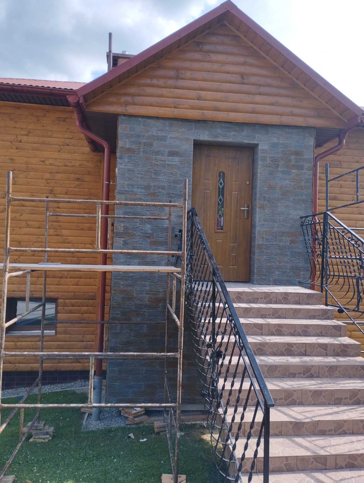 Dom z drewnianą elewacją, fragment ściany wykończony szarym kamieniem dekoracyjnym, wejście z brązowymi drzwiami i schodami wyłożonymi płytkami, widoczne rusztowanie.