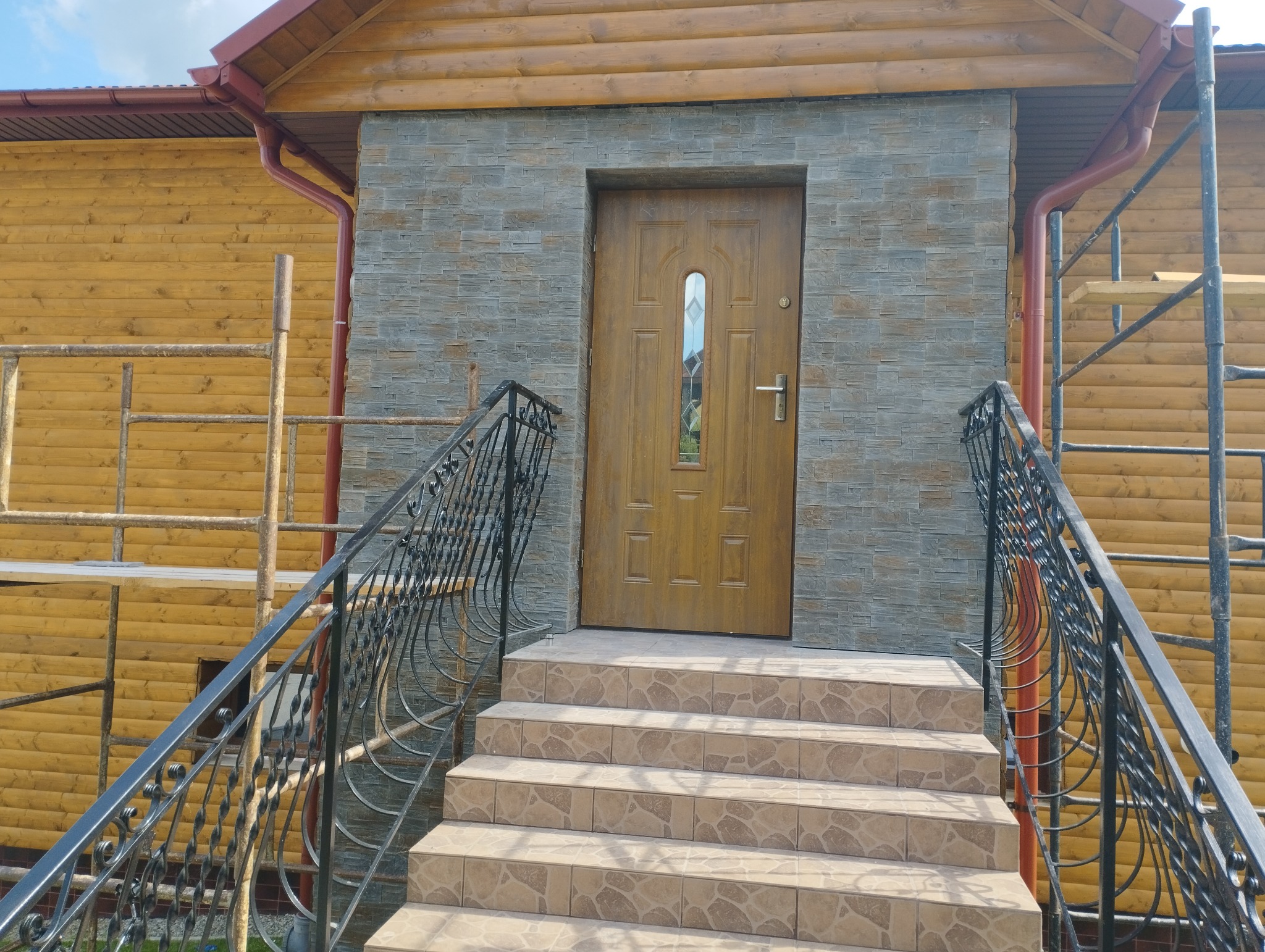 Wejście do drewnianego domu z elewacją wykończoną szarą, kamienną okładziną, brązowe drzwi z szybką, schody wyłożone beżowymi płytkami z wzorem, czarne, ozdobne balustrady, rusztowanie z boku.