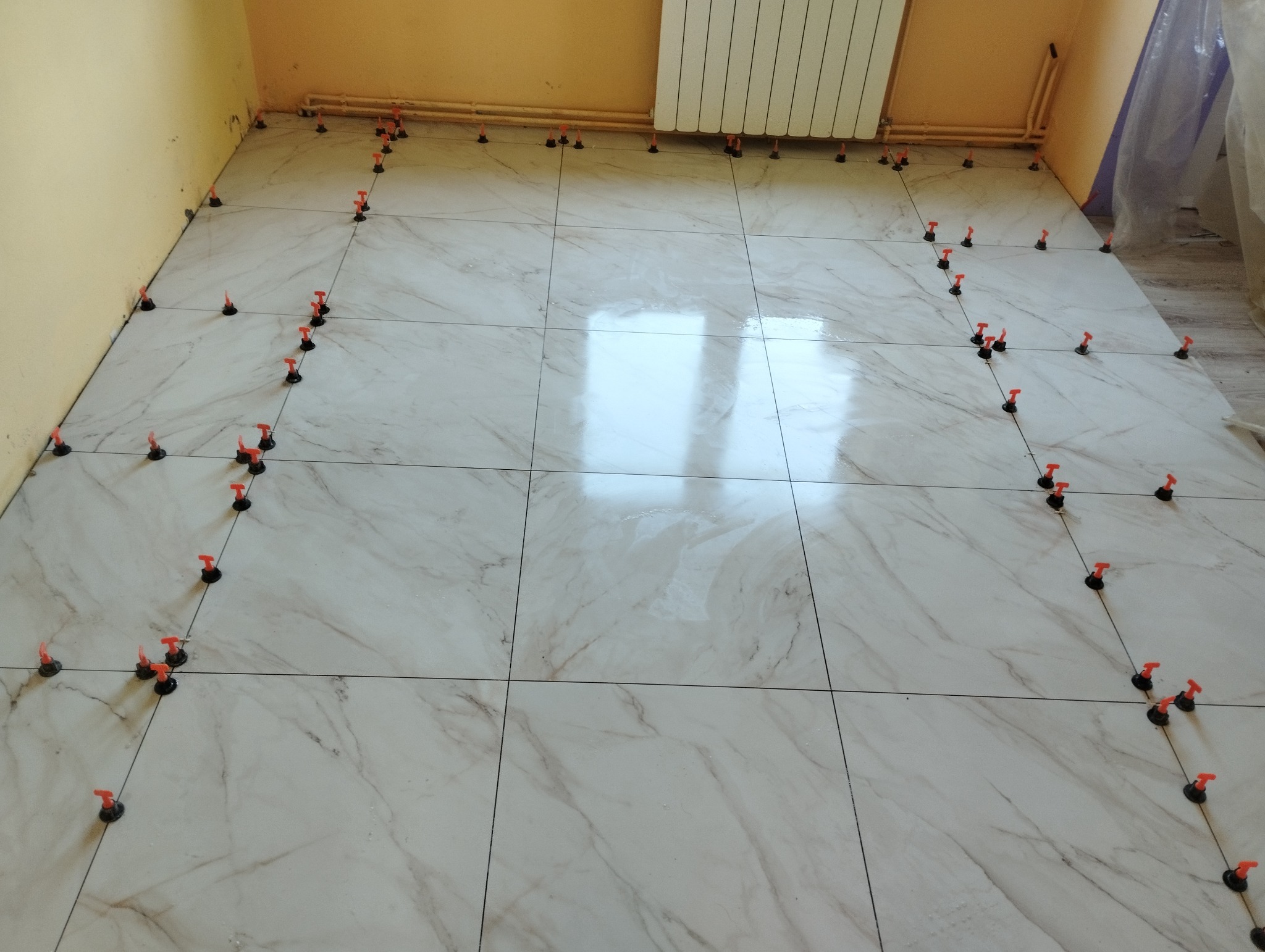 Układanie płytek podłogowych imitujących marmur, z użyciem systemu poziomowania z czerwonymi klinami, widoczne niedoskonałości w narożniku i przy rurach grzejnika.