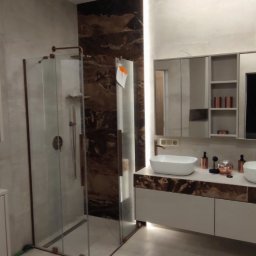 Matrix bud - Nowoczesna łazienka z kabiną walk-in, szklanymi drzwiami i miedzianymi akcentami. Podwójna umywalka na szafce z ciemnym blatem, jasne płytki podłogowe i ścienne, oświetlenie LED.