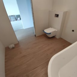Matrix bud - Nowocześnie wykończona łazienka z podwieszaną toaletą w ochronnej folii, jasną podłogą imitującą drewno i białymi płytkami ściennymi. Widoczna część wanny.