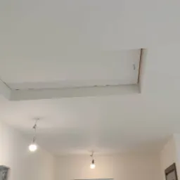 Biały sufit z zamontowaną płytą kartonowo-gipsową, tworzącą otw&oacute;r rewizyjny z widocznym metalowym uchwytem, oświetlony przez trzy wiszące żar&oacute;wki na długich kablach.